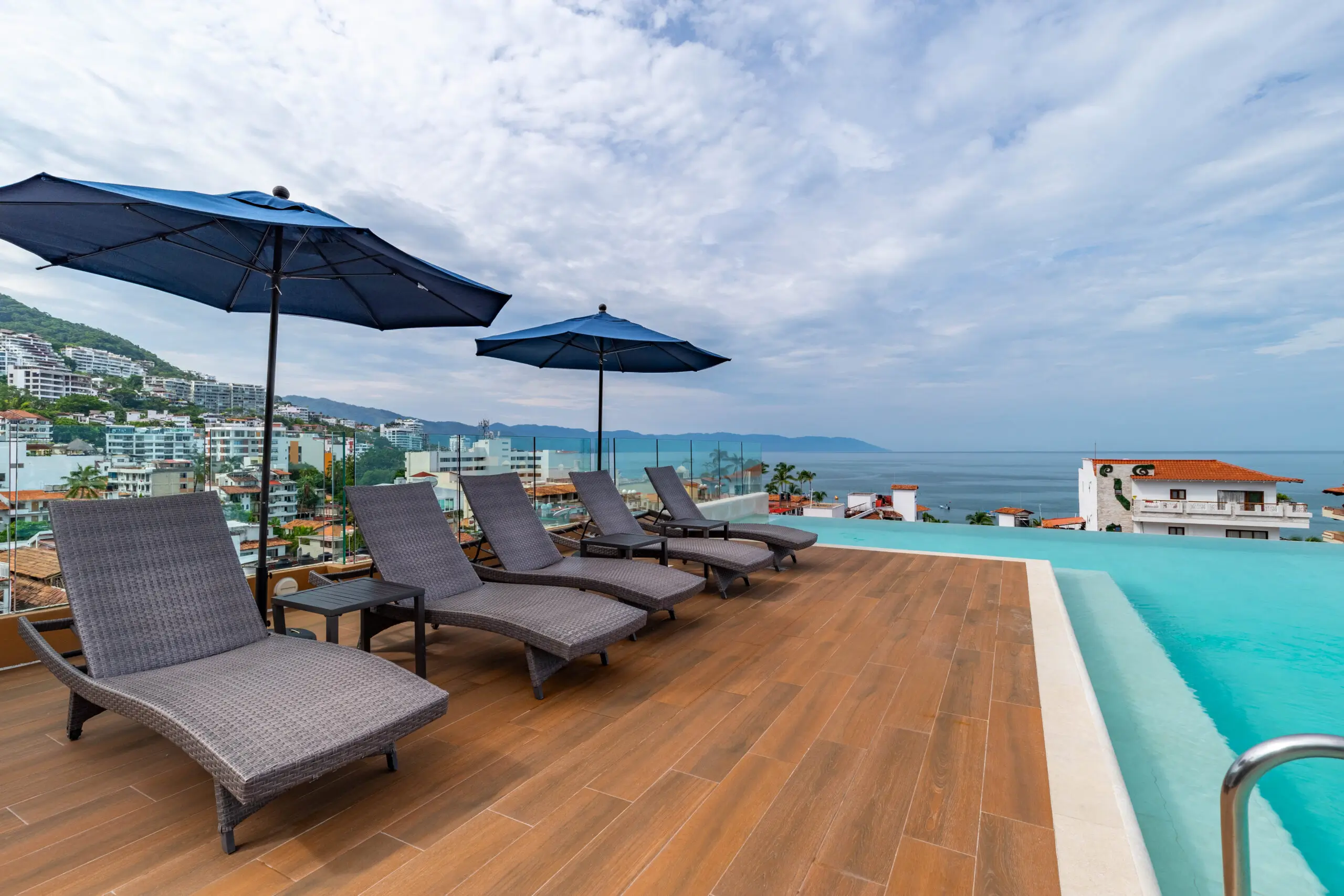 Poolside lounge chairs with umbrellas overlooking the ocean under a cloudy sky.